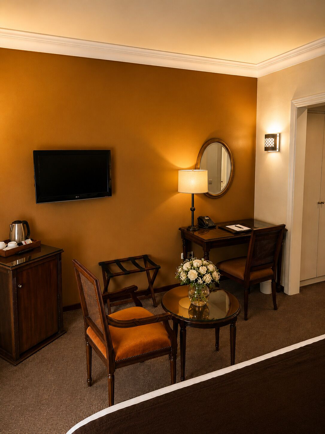 A hotel room with a flat-screen TV, a desk with a lamp, a chair, a small table with a vase of flowers, and a cabinet.