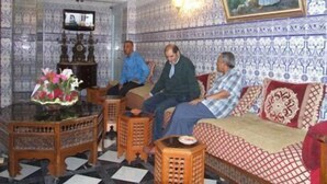 Una habitación con paredes estampadas, un sofá de madera, una mesita con flores y un televisor sobre un mueble.