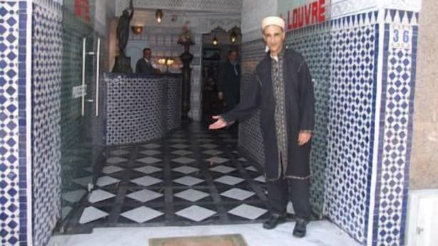 Un pasillo con paredes de baldosas estampadas y suelo a cuadros, que conduce a una zona de recepción con mostrador y un letrero que dice "L'LOUVRE".