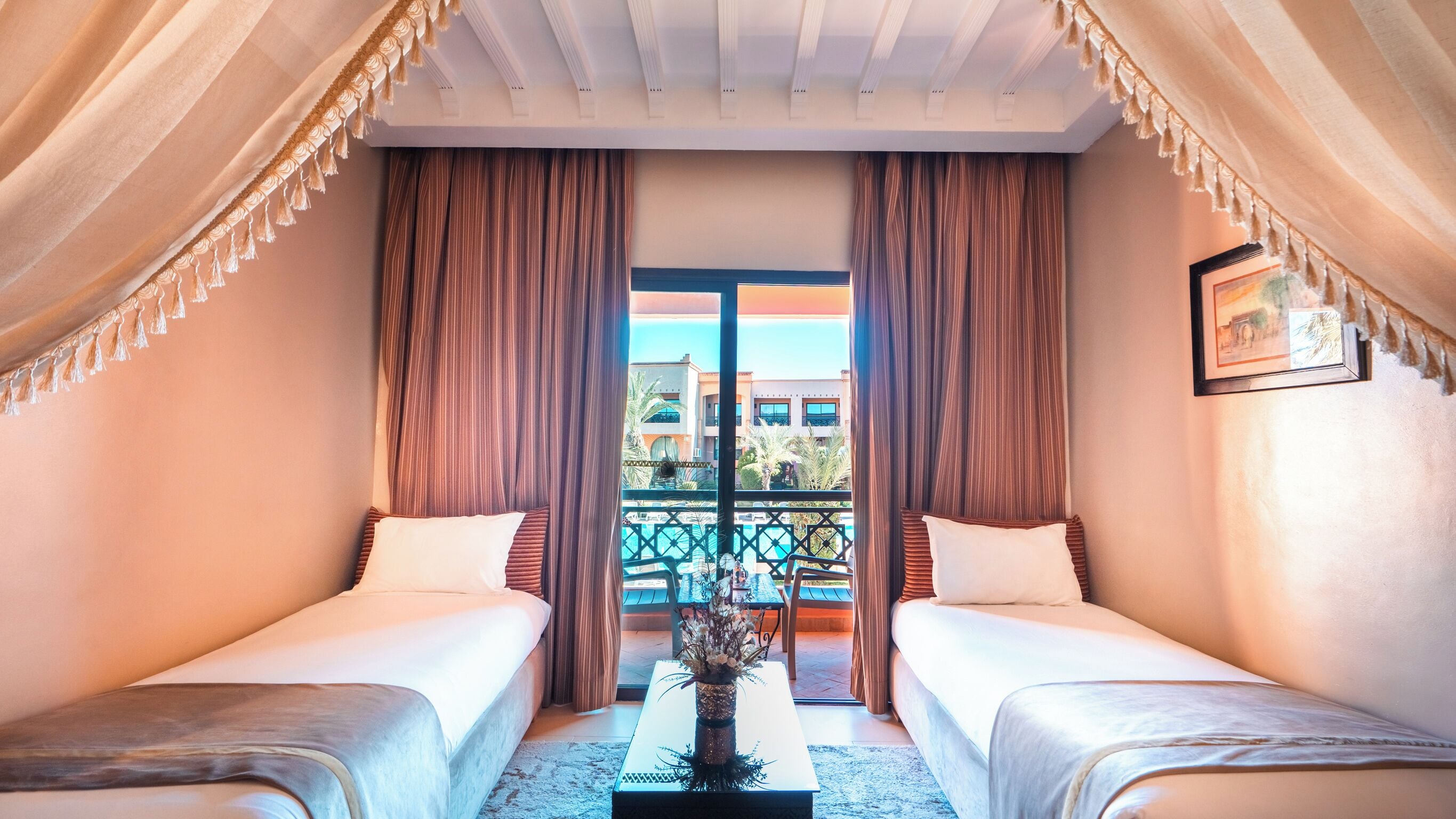 Une chambre d’hôtel avec deux lits, un balcon donnant sur des immeubles et une table agrémentée d’un vase de fleurs.