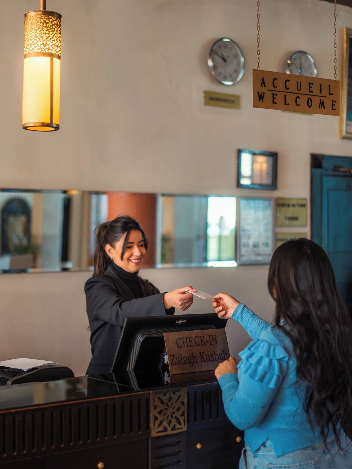 A hotel reception area with a check-in counter, a 'WELCOME' sign, and a clock on the wall.