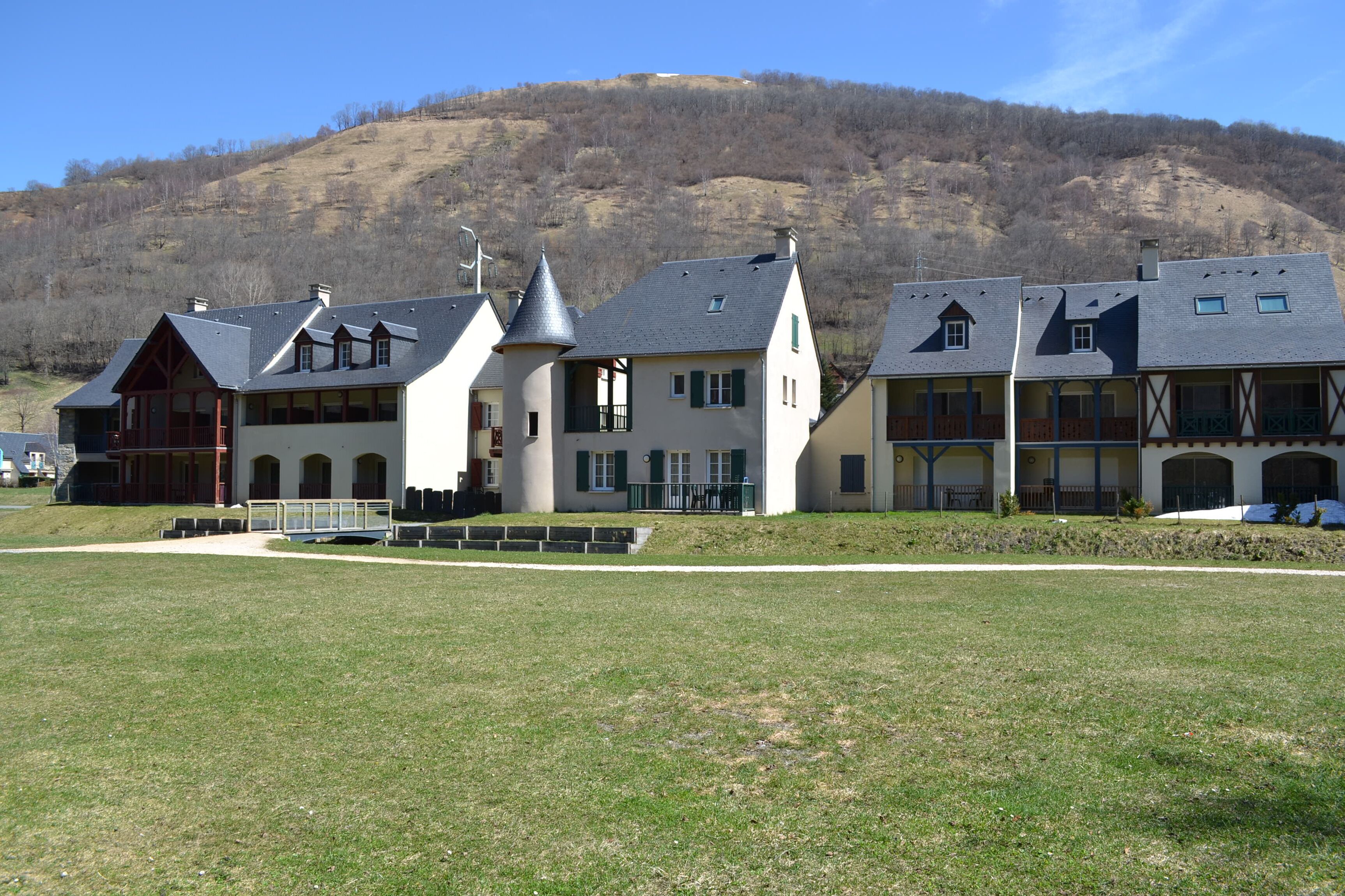 Uma fileira de edifícios tradicionais em estilo europeu, com um campo gramado em primeiro plano e uma colina ao fundo.