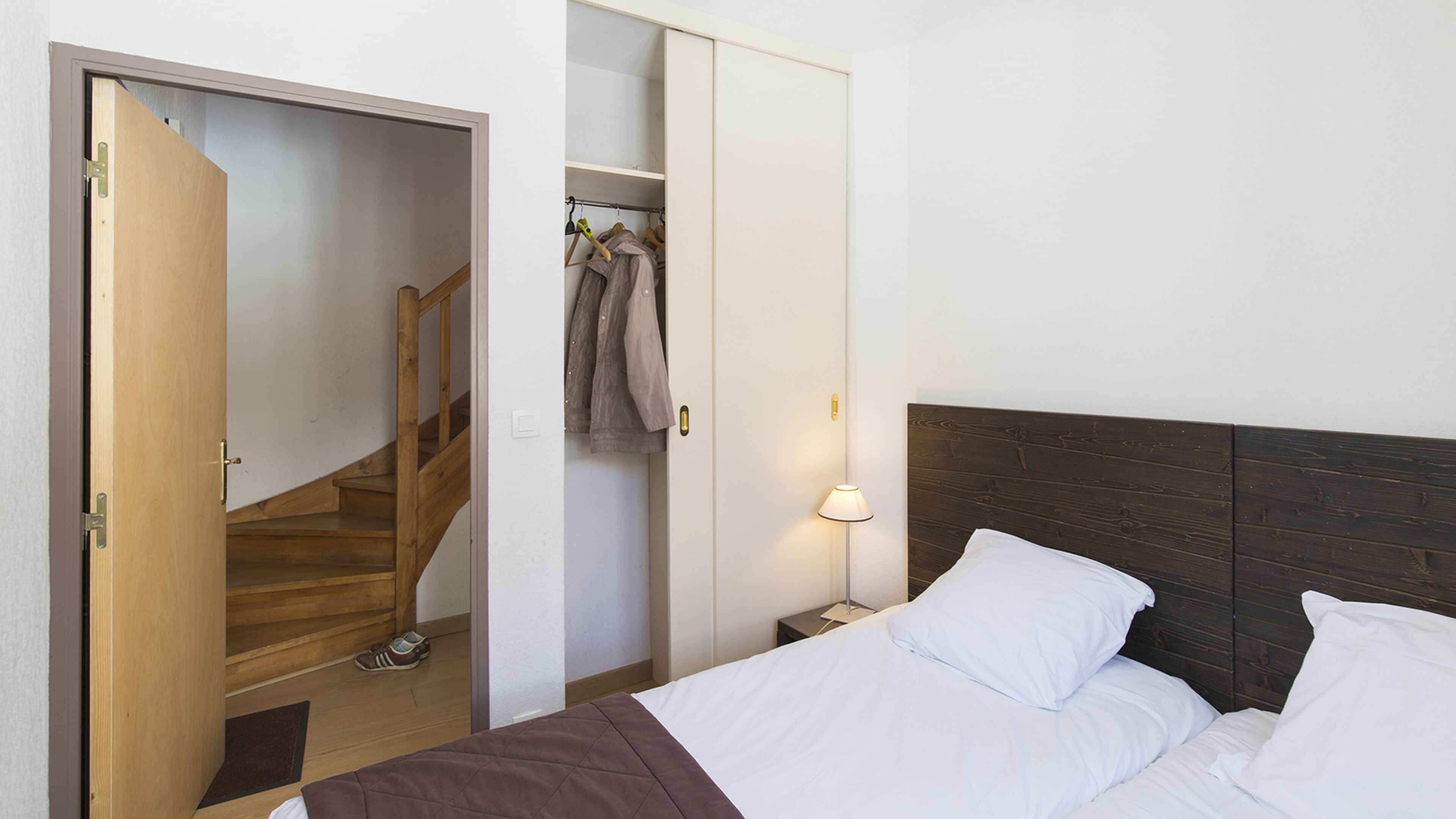 A bedroom with a wooden staircase, a closet with hanging clothes, a bedside table with a lamp, and a bed with white bedding.