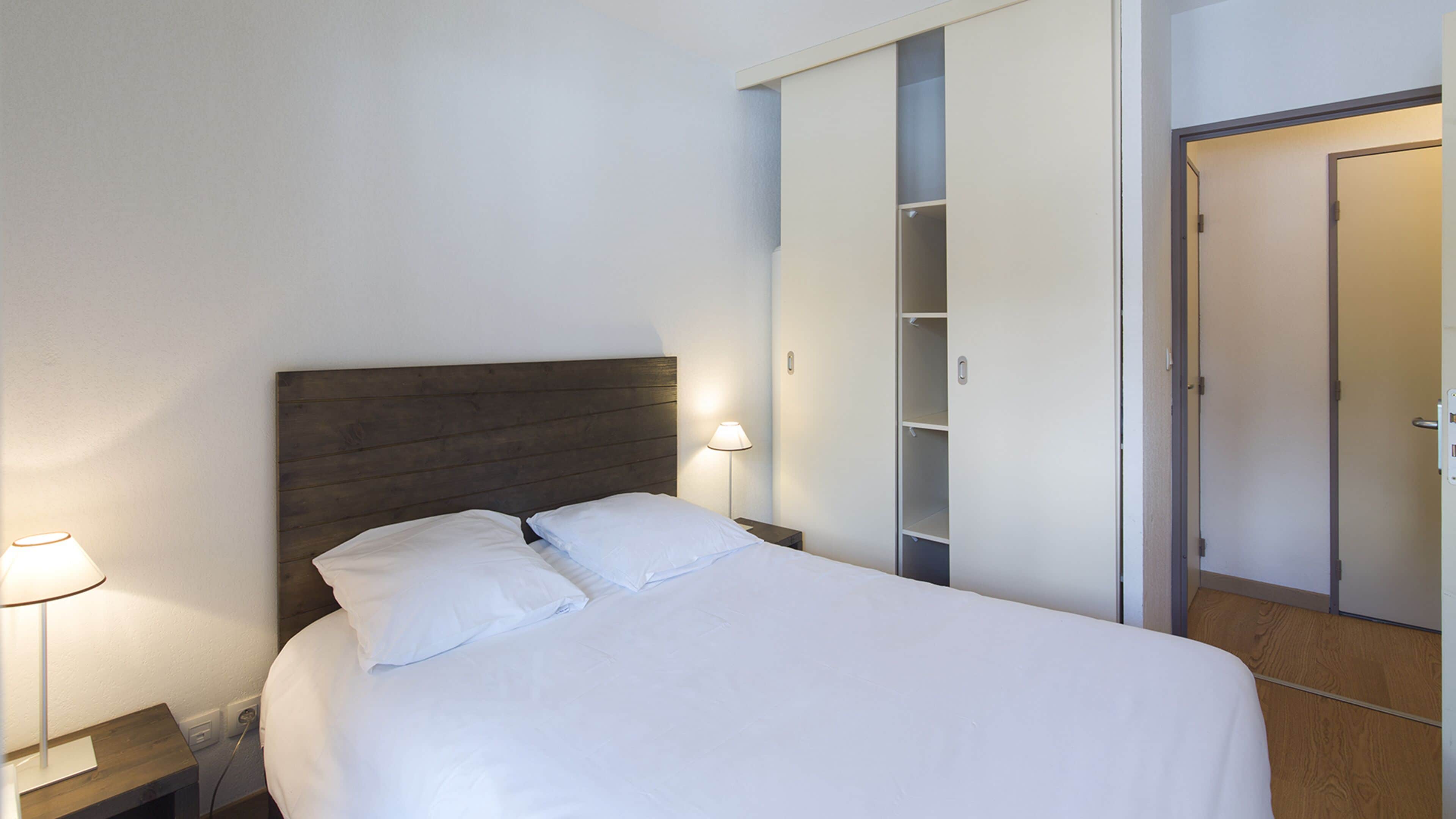 A neatly made bed with white linens in a hotel room. There is a wooden headboard, a bedside lamp, and a wardrobe with sliding doors.