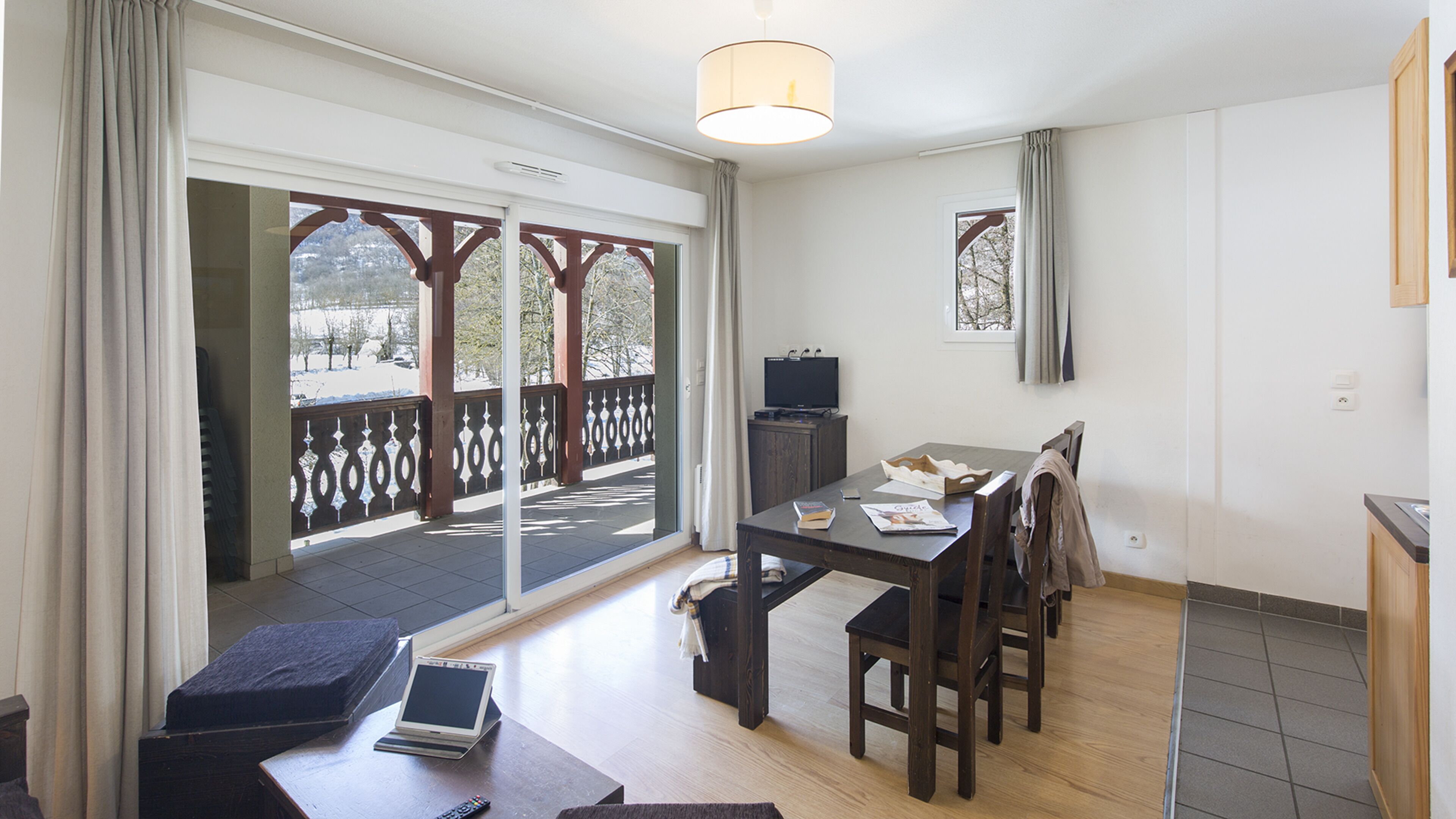Een moderne woonkamer met een houten eettafel, een bank en een televisie. Er is een balkon met uitzicht op bomen en een besneeerd landschap.
