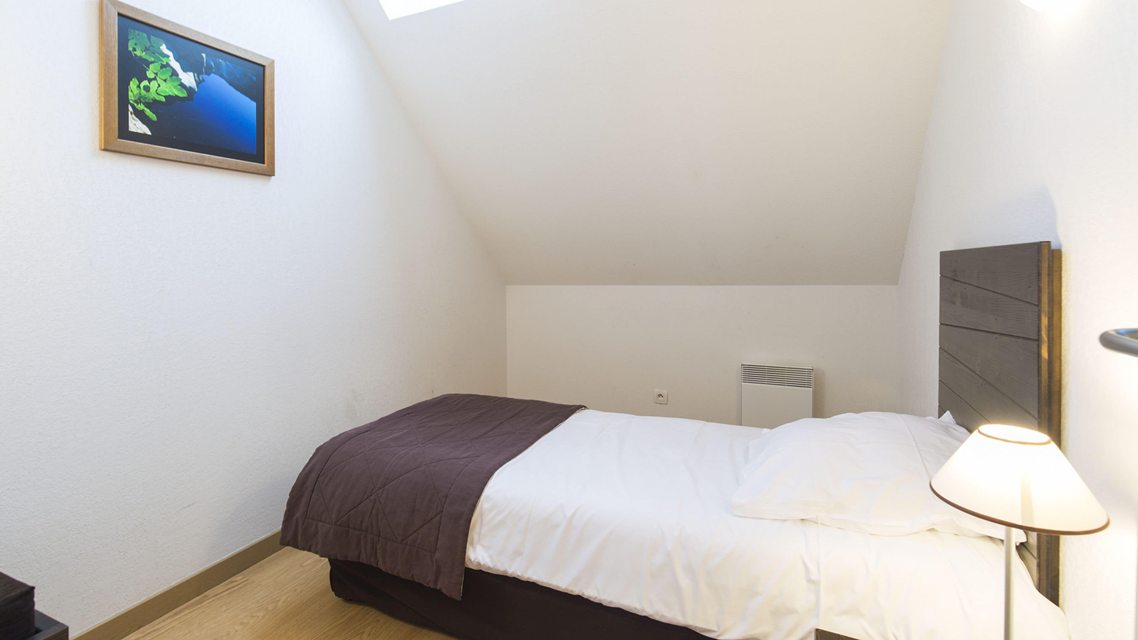 Un dormitorio pequeño y ordenado con una cama individual, una mesita de noche con lámpara, un radiador y un cuadro en la pared.