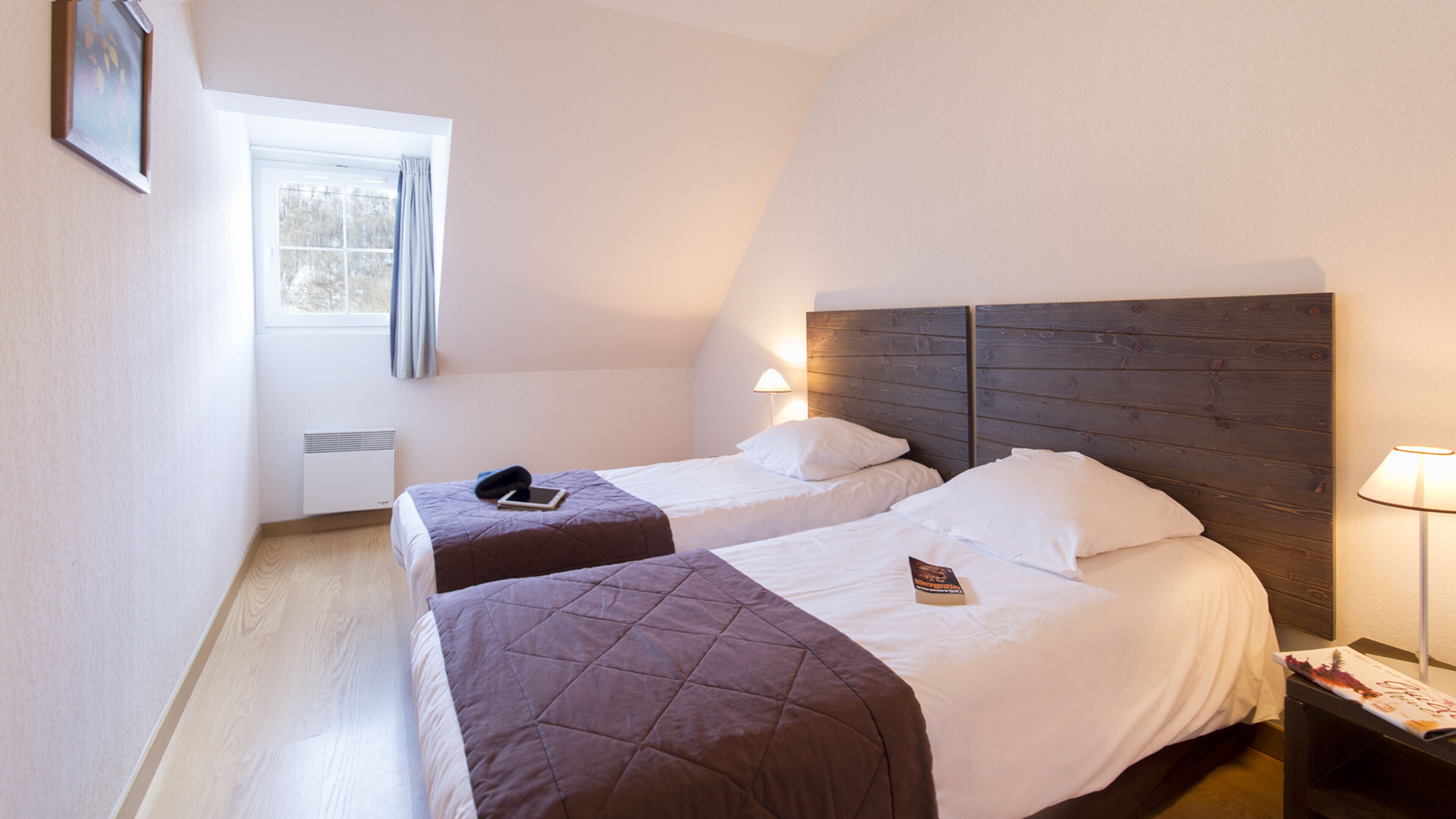 A hotel room with two single beds, a window with curtains, a bedside table with a lamp, and a framed picture on the wall.
