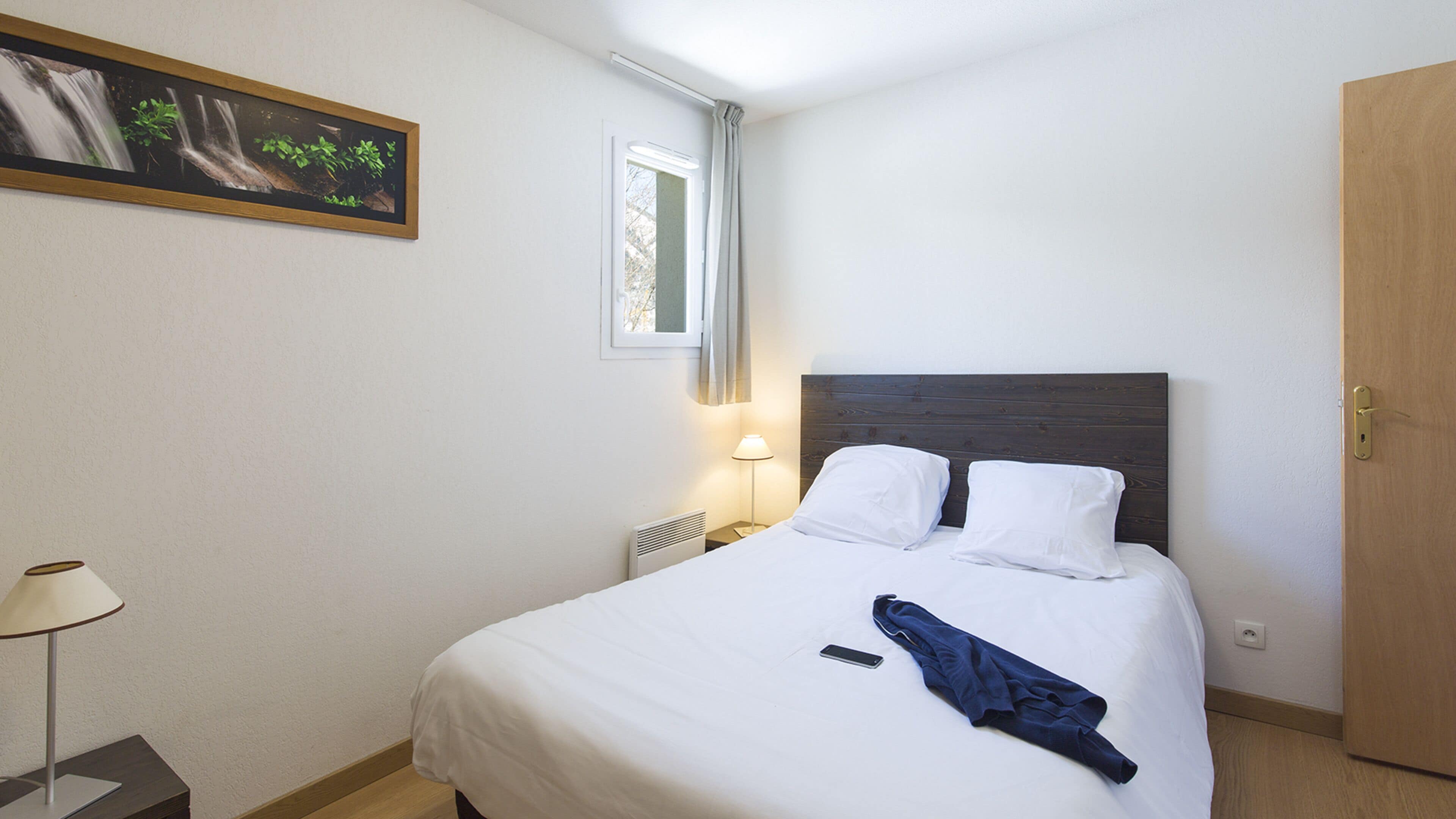 A neatly made bed with white linens, a bedside lamp, and a smartphone on the bed.