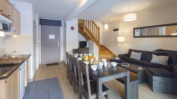 A modern kitchen with a microwave, sink, and wooden cabinets. There is a dining area with a table set for four, a staircase, and a living room with a sofa and a TV.