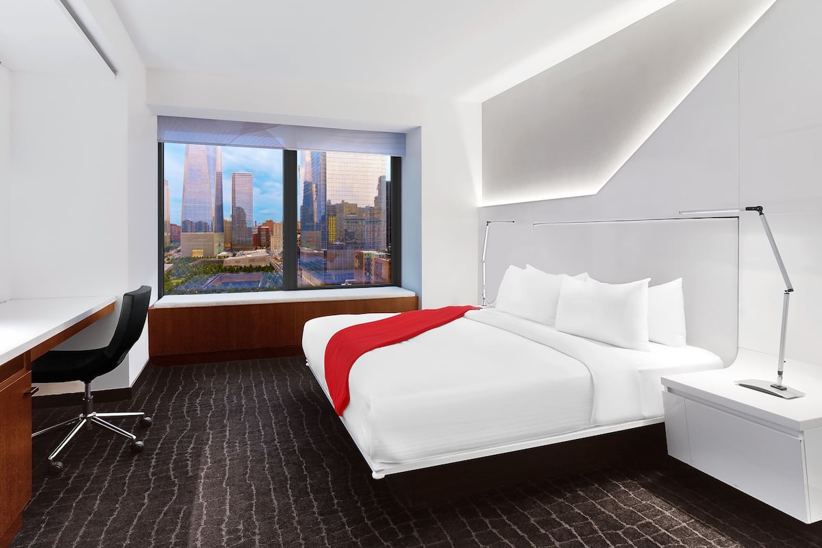 A modern hotel room with a city view, a desk, and a bed with white bedding.