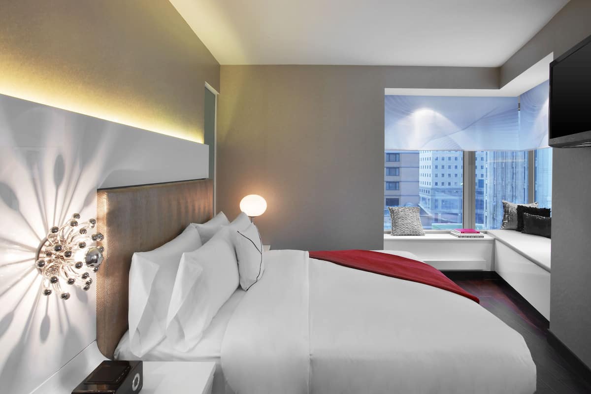 A modern hotel room with a large bed, a bedside table, a lamp, and a window with a city view.