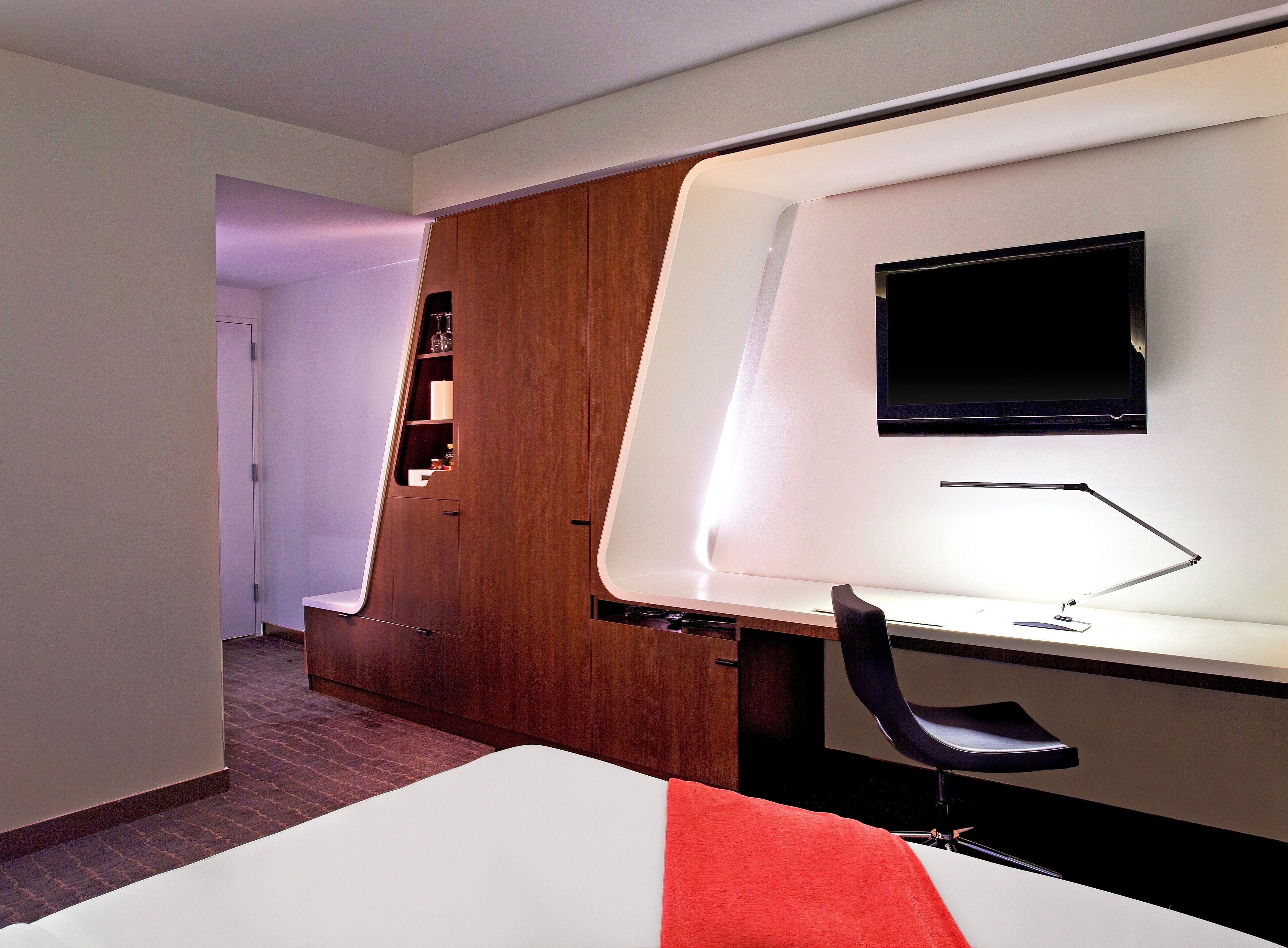Une chambre d’hôtel moderne équipée d’un lit, d’un bureau avec une chaise, d’un téléviseur fixé au mur et d’une armoire en bois.