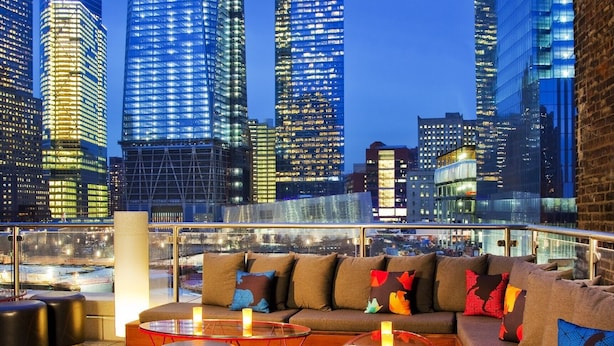 Un salón en la azotea con vistas a un horizonte urbano, que cuenta con una zona de estar moderna con sofás y mesas, y una barandilla de vidrio con vistas a la ciudad.