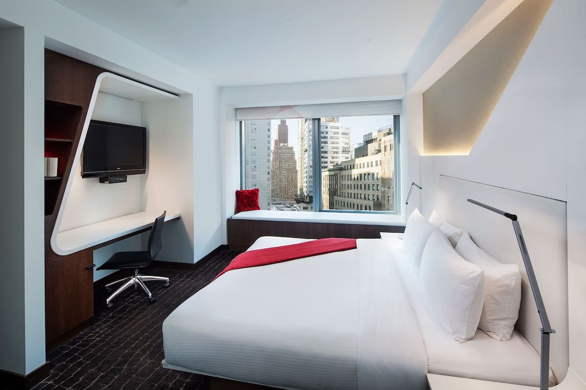 A modern hotel room with a large bed, a desk, a chair, a TV, and a window with a city view.