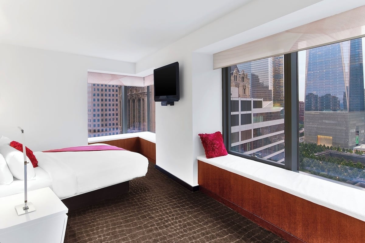A hotel room with a large window offering a city view, a bed with white bedding and red pillows, a flat-screen TV mounted on the wall, and a small white side table.