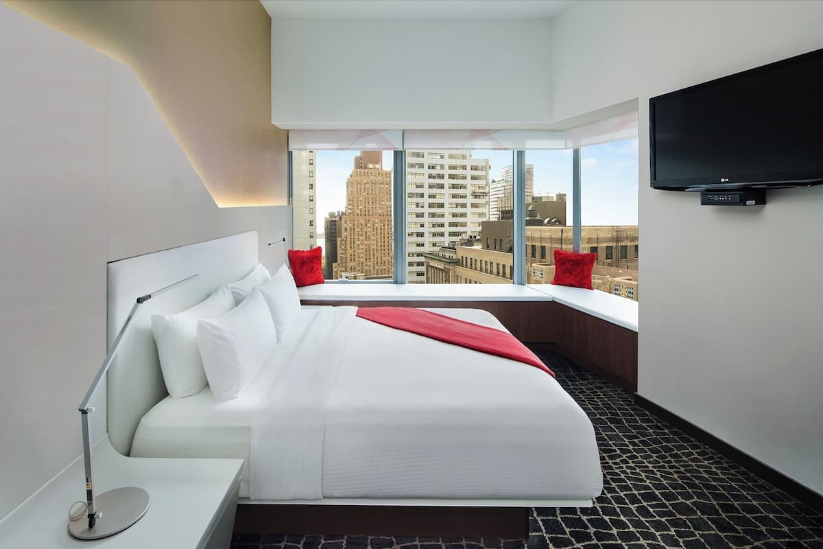A modern hotel room with a large bed, a desk, a desk lamp, a flat-screen TV, and a city view through large windows.