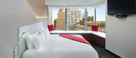 A modern hotel room with a large bed, a desk, a desk lamp, a flat-screen TV, and a city view through large windows.