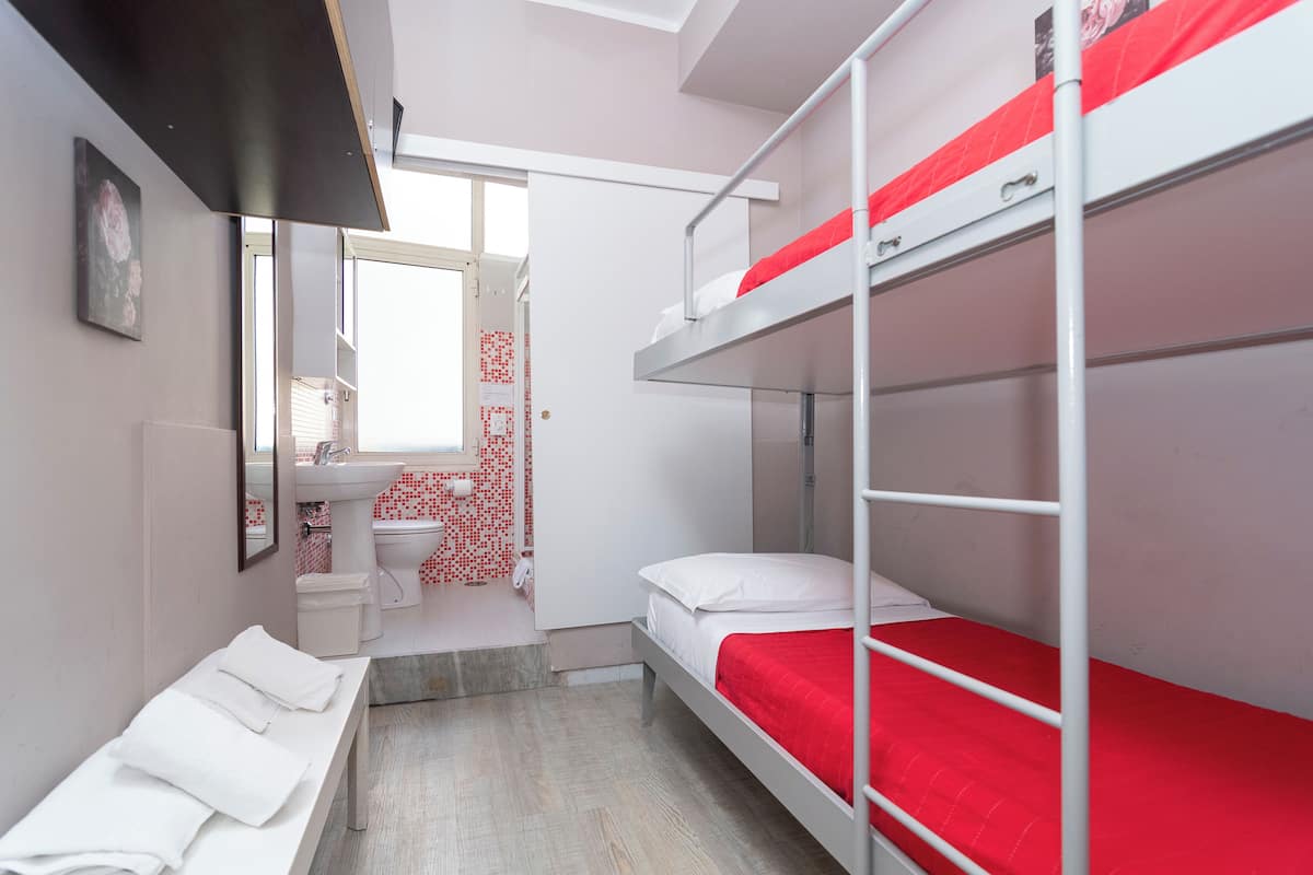 Ein Stockbett mit einer roten Matratze, eine weiße Bank mit Handtüchern, ein Badezimmer mit rot-weißen Fliesen und ein Fenster mit Blick.