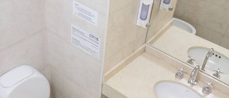 A bathroom with a toilet, sink, and mirror. There is a sign on the wall and a trash bin.