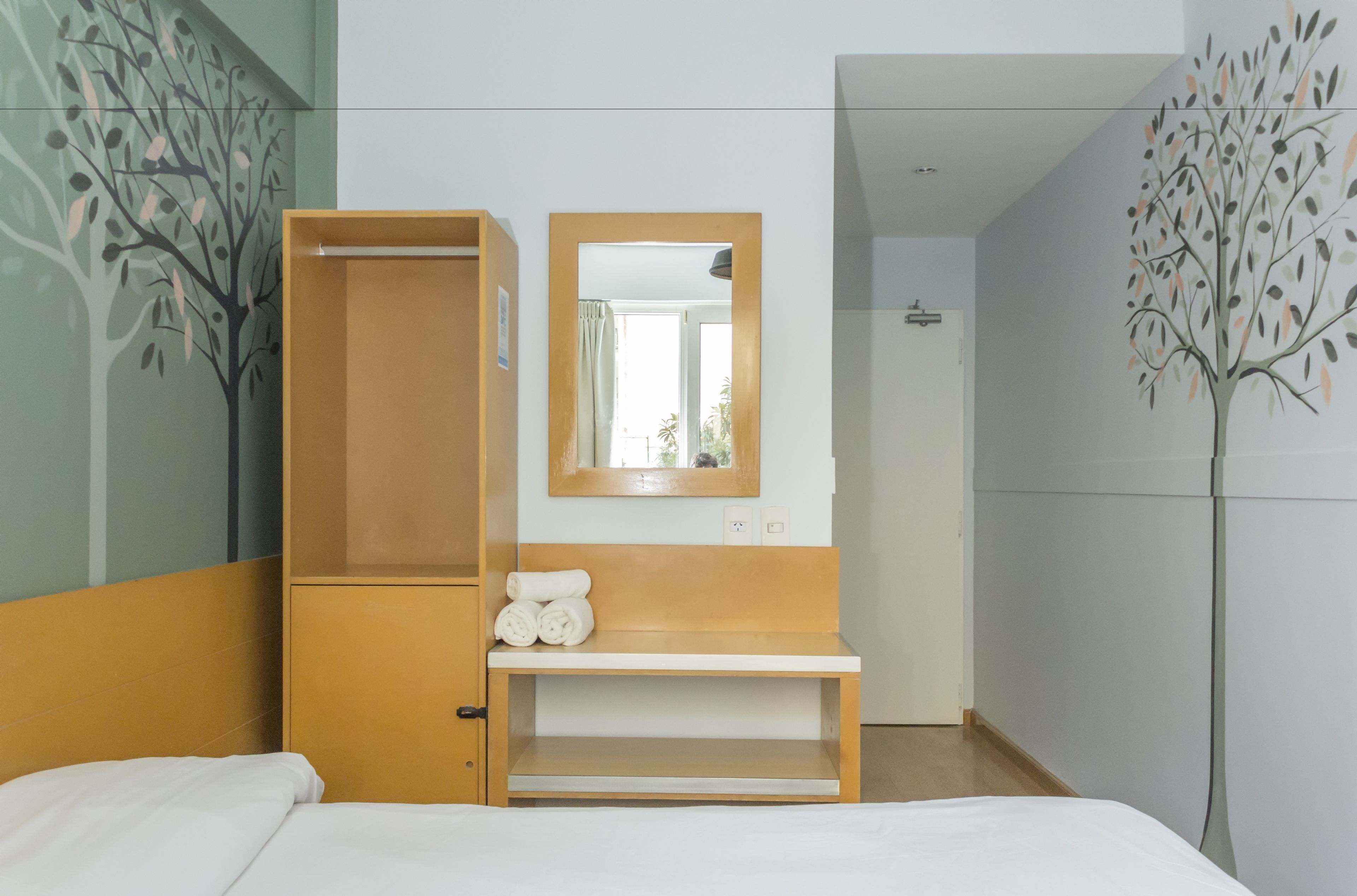Une chambre d’hôtel avec un lit, une armoire en bois, un miroir et une étagère fixée au mur où sont rangées les serviettes.