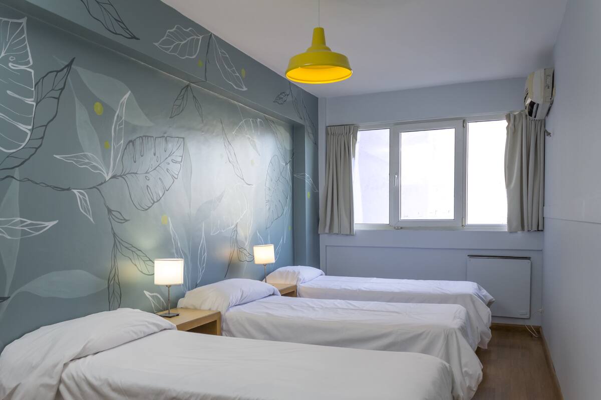Quarto com duas camas, um mural na parede, uma janela com cortinas e uma luminária pendente amarela.
