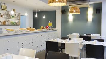 A modern dining area with white tables and black chairs, a counter with food and drinks, and wicker pendant lights.