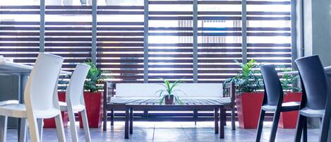 A modern indoor seating area with a long table and chairs, potted plants, and a view through large windows with horizontal blinds.