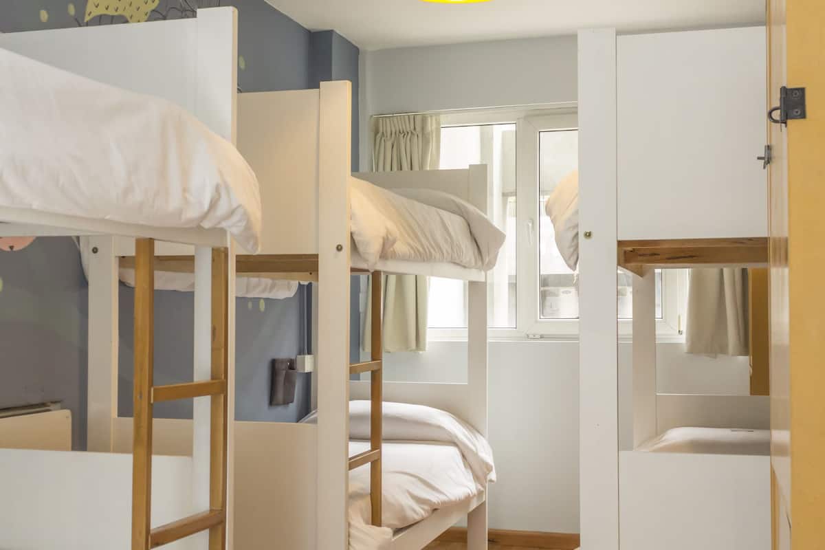 Quarto com beliches, piso de madeira, luminária pendente amarela e janela com cortinas.