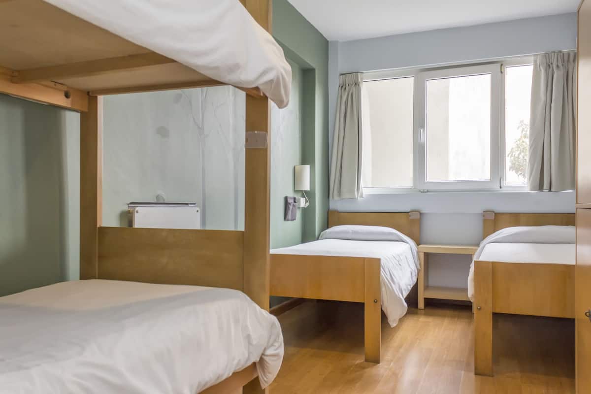 Um quarto com três beliches de madeira, uma pequena mesa de cabeceira com abajur e uma janela com cortinas.