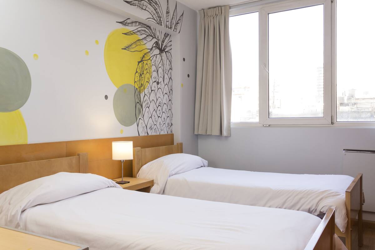 Quarto de hotel com duas camas, uma mesa de cabeceira com abajur, um mural na parede e uma janela com cortinas.
