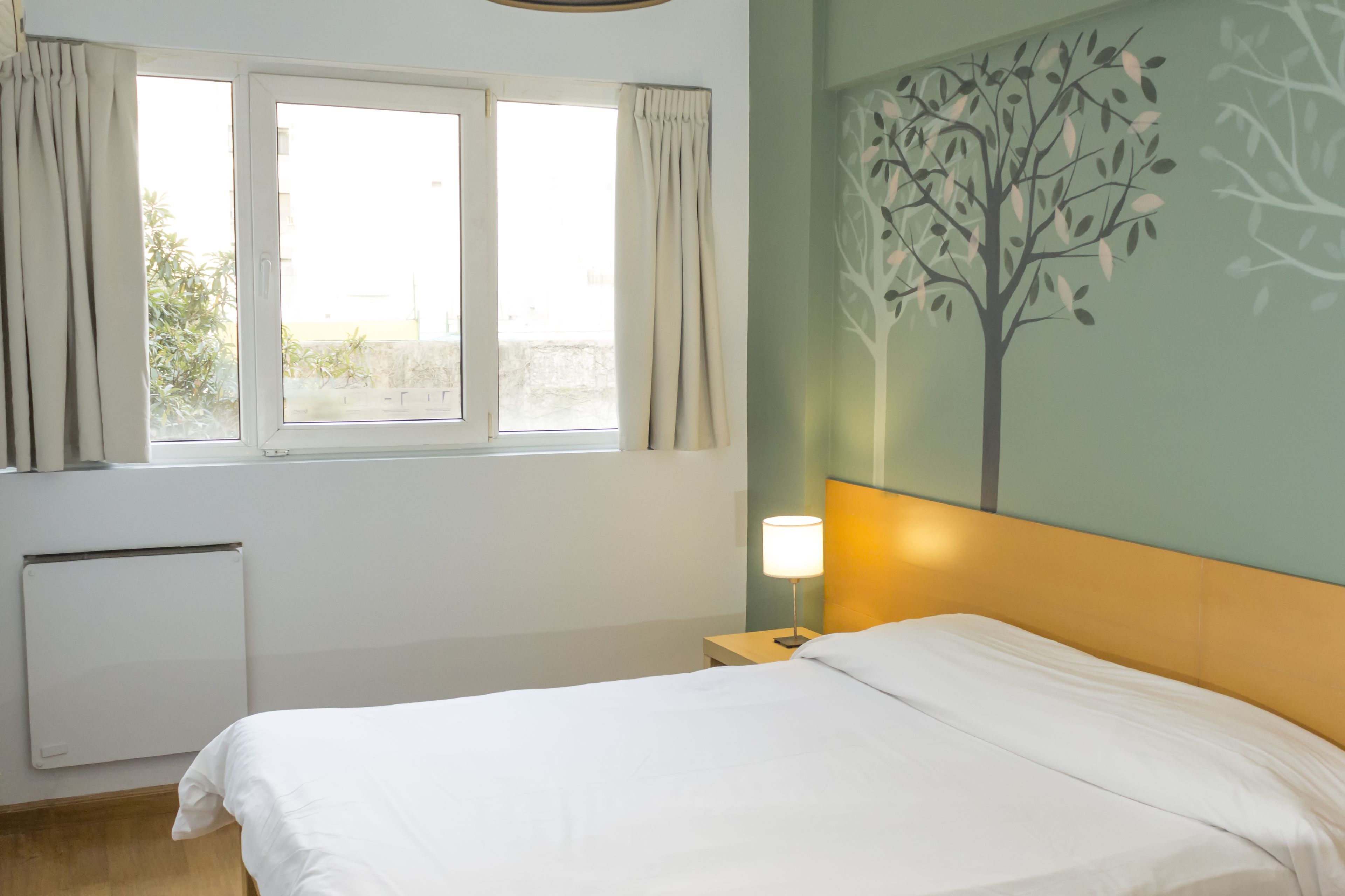 A hotel room with a bed, a nightstand, a lamp, a radiator, and a window with curtains.