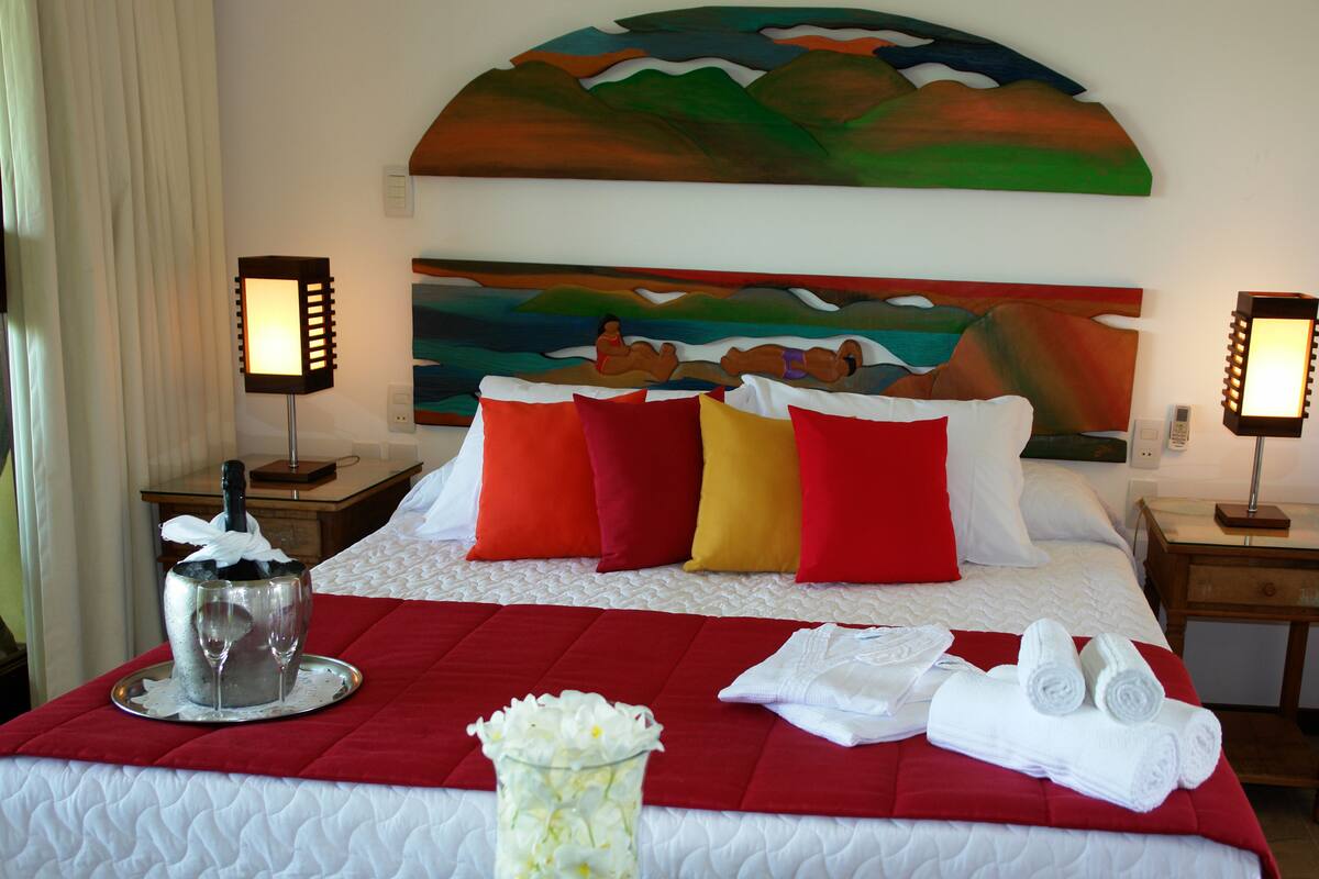 Une chambre d’hôtel avec un lit, des oreillers rouges et blancs, un plateau avec une bouteille et des verres, un vase avec des fleurs et un tableau au mur.