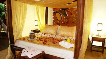 Una cama con dosel y cuatro columnas, una mesita de noche con lámpara, y una pequeña mesa con un arreglo floral.