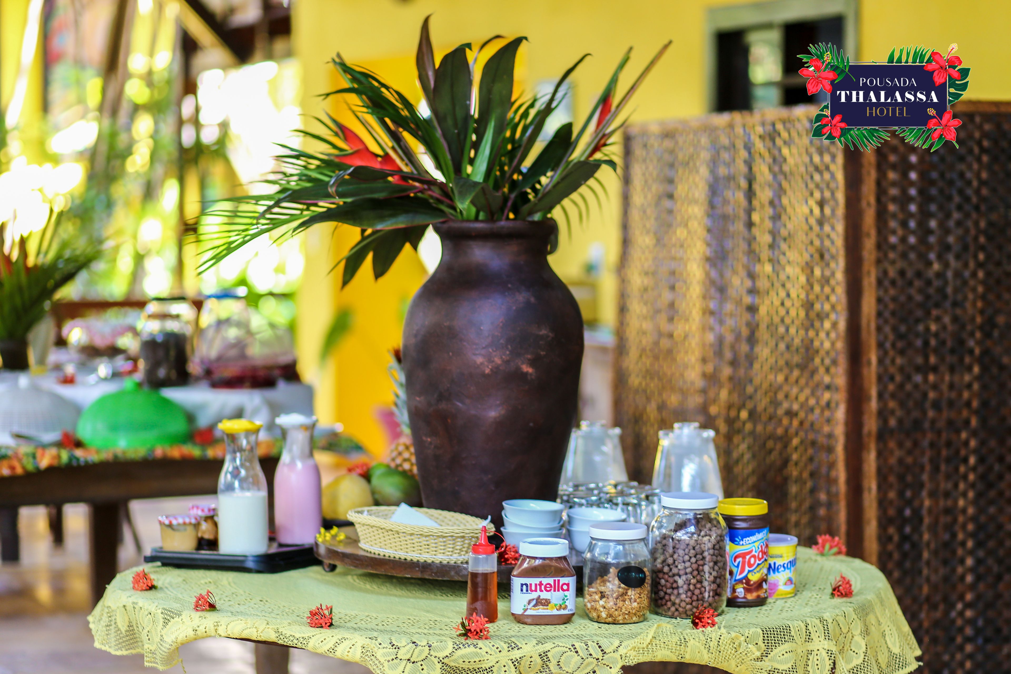 A table with various food items and condiments, a large vase with green plants, and a sign for Pousada Thalassa Hotel.