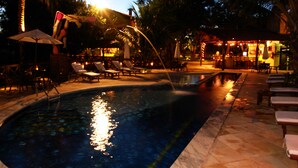 Une piscine entourée de palmiers, avec des chaises longues et des parasols, ainsi qu’un espace bar.