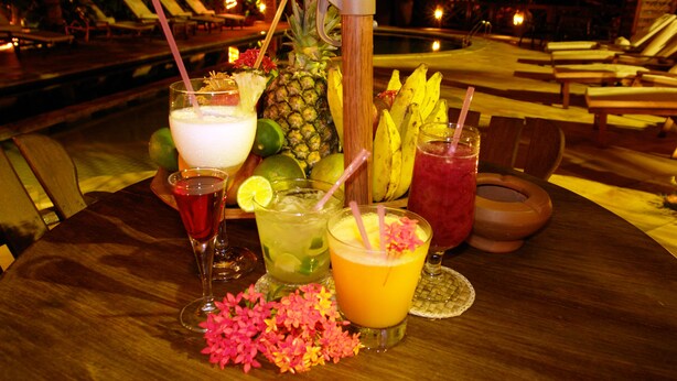 Une table garnie de diverses boissons et de fruits tropicaux, entourée de fauteuils et avec une piscine en arrière-plan.