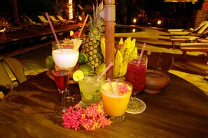 A table with various drinks and tropical fruits, surrounded by lounge chairs and a pool area in the background.