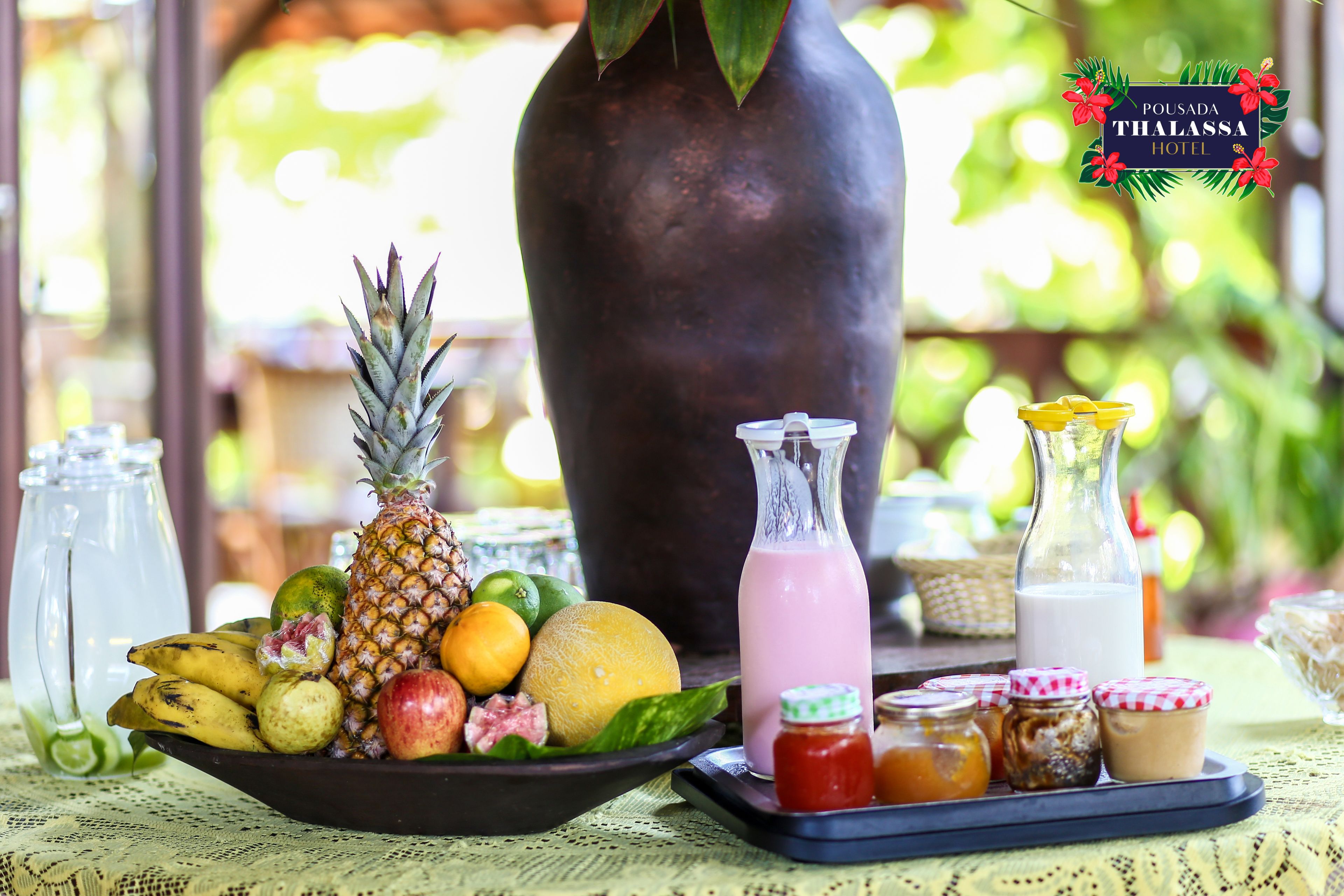 A tropical fruit bowl with a bottle of pink drink, jars of condiments, and a large vase on a table.