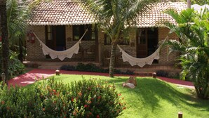 Une maison à deux étages, avec un toit en tuiles, un hamac et un jardin bien entretenu agrémenté de fleurs rouges et de palmiers.