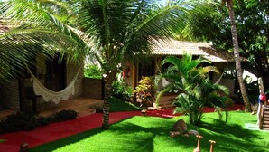 Un jardin luxuriant avec un hamac, des palmiers et un chemin rouge menant à un bâtiment doté d’un toit en tuiles.