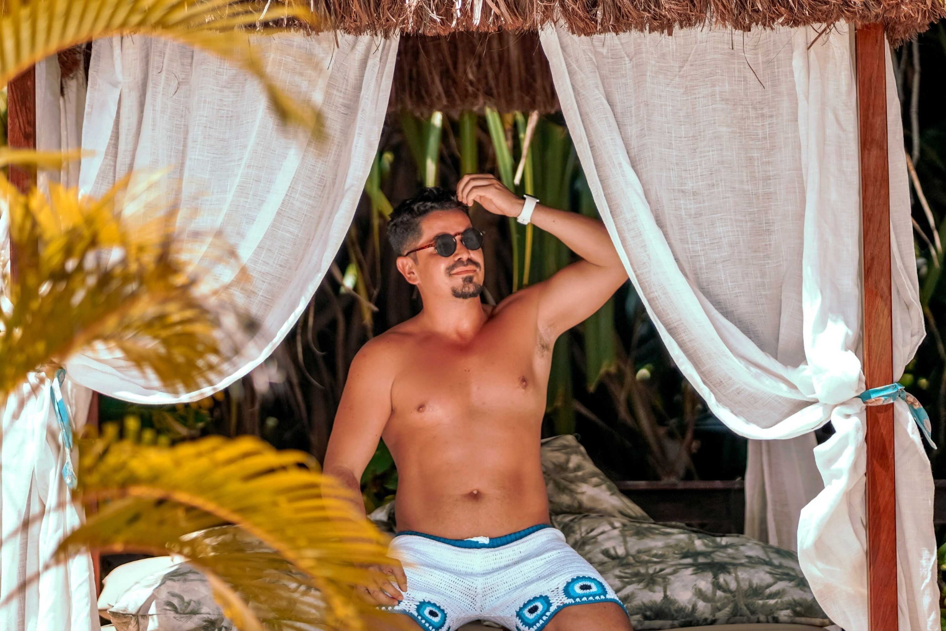 A man in a hammock under a thatched roof, surrounded by tropical plants.