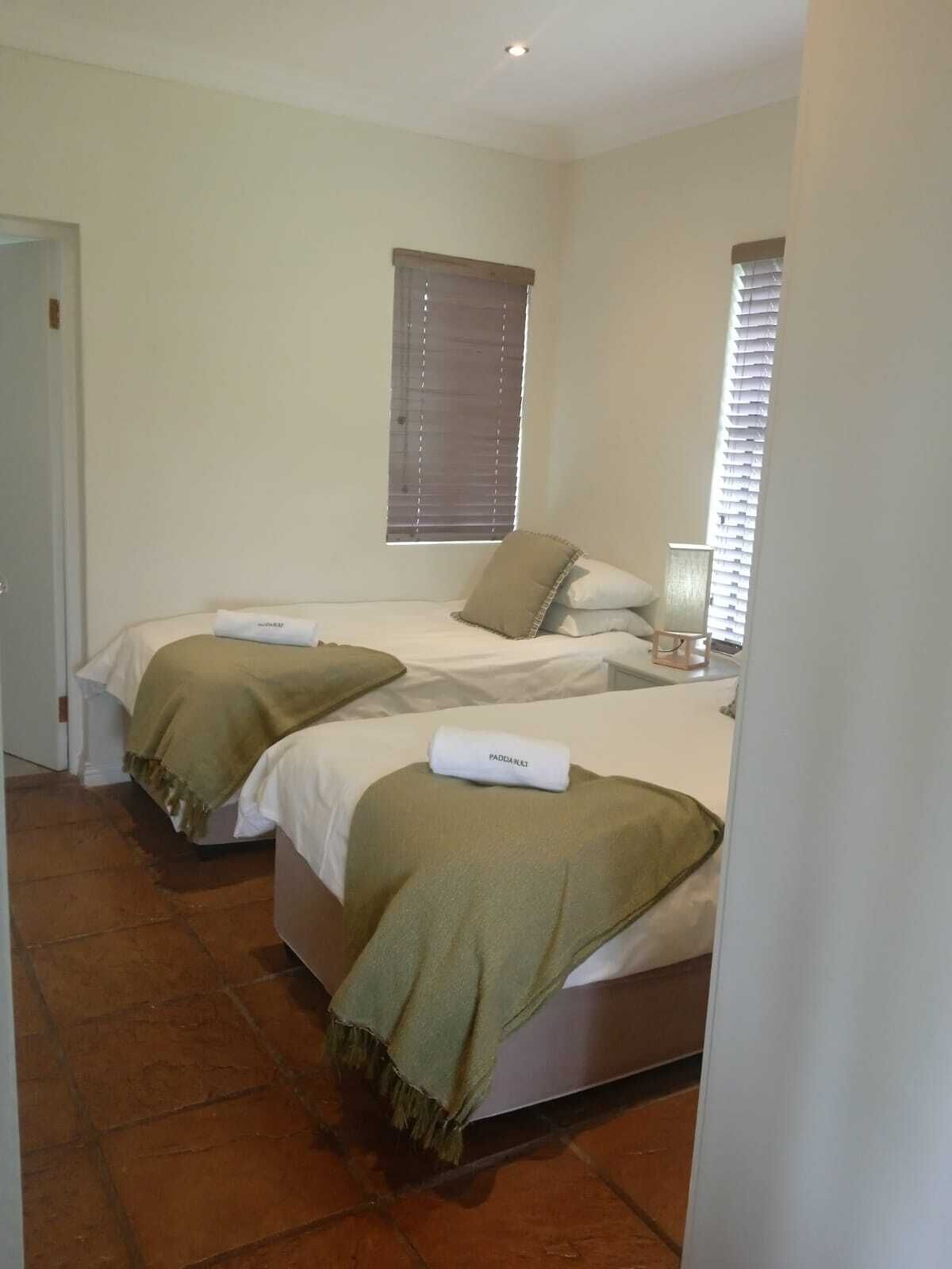 A hotel room with two beds, each with a pillow and a towel. There is a window with blinds, a door, and a small table with a lamp.