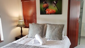 Una cama bien hecha con una colcha a rayas, dos almohadas y una toalla enrollada. Hay una lámpara en una mesita de noche, un cuadro de pájaros en la pared y una puerta a la derecha.