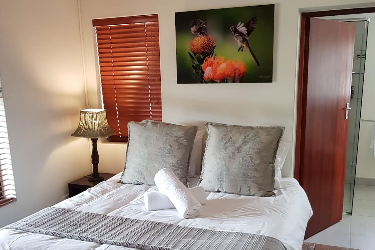 A neatly made bed with a striped bedspread, two pillows, and a rolled towel. There is a lamp on a bedside table, a painting of birds on the wall, and a door to the right.