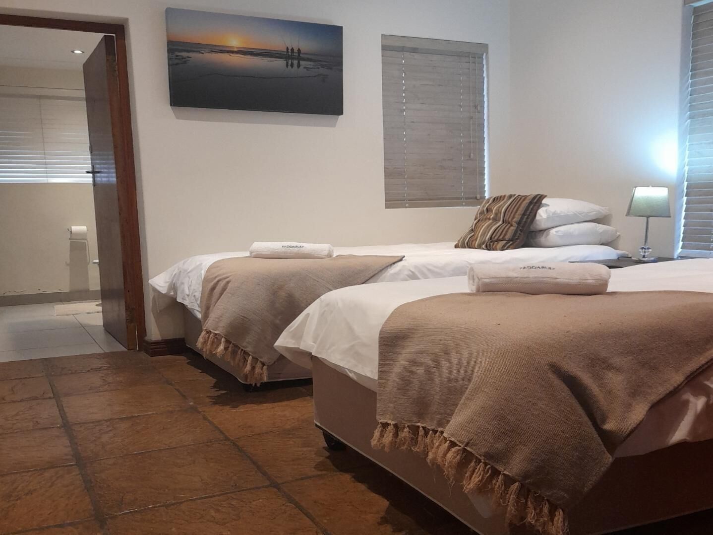 Una habitación de hotel con dos camas, un cuadro de un atardecer y una ventana con persianas.