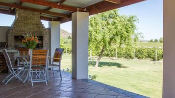 A covered patio with a stone fireplace, outdoor seating, and a view of a grassy landscape.
