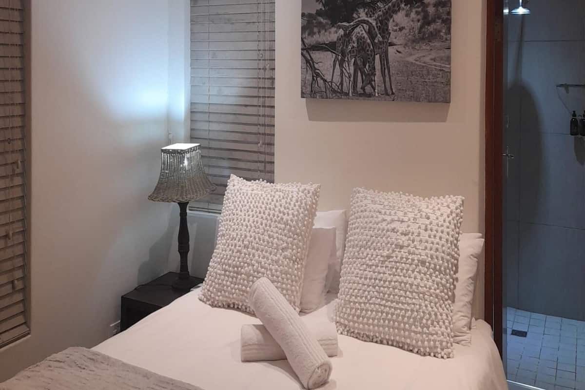 A bedroom with a bed, two pillows, a rolled towel, a nightstand with a lamp, and a painting on the wall.