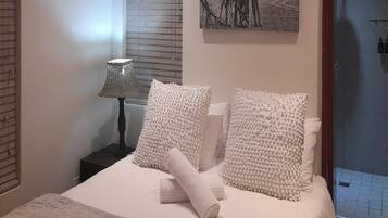 Un dormitorio con una cama, dos almohadas, una toalla enrollada, una mesita de noche con una lámpara y un cuadro en la pared.
