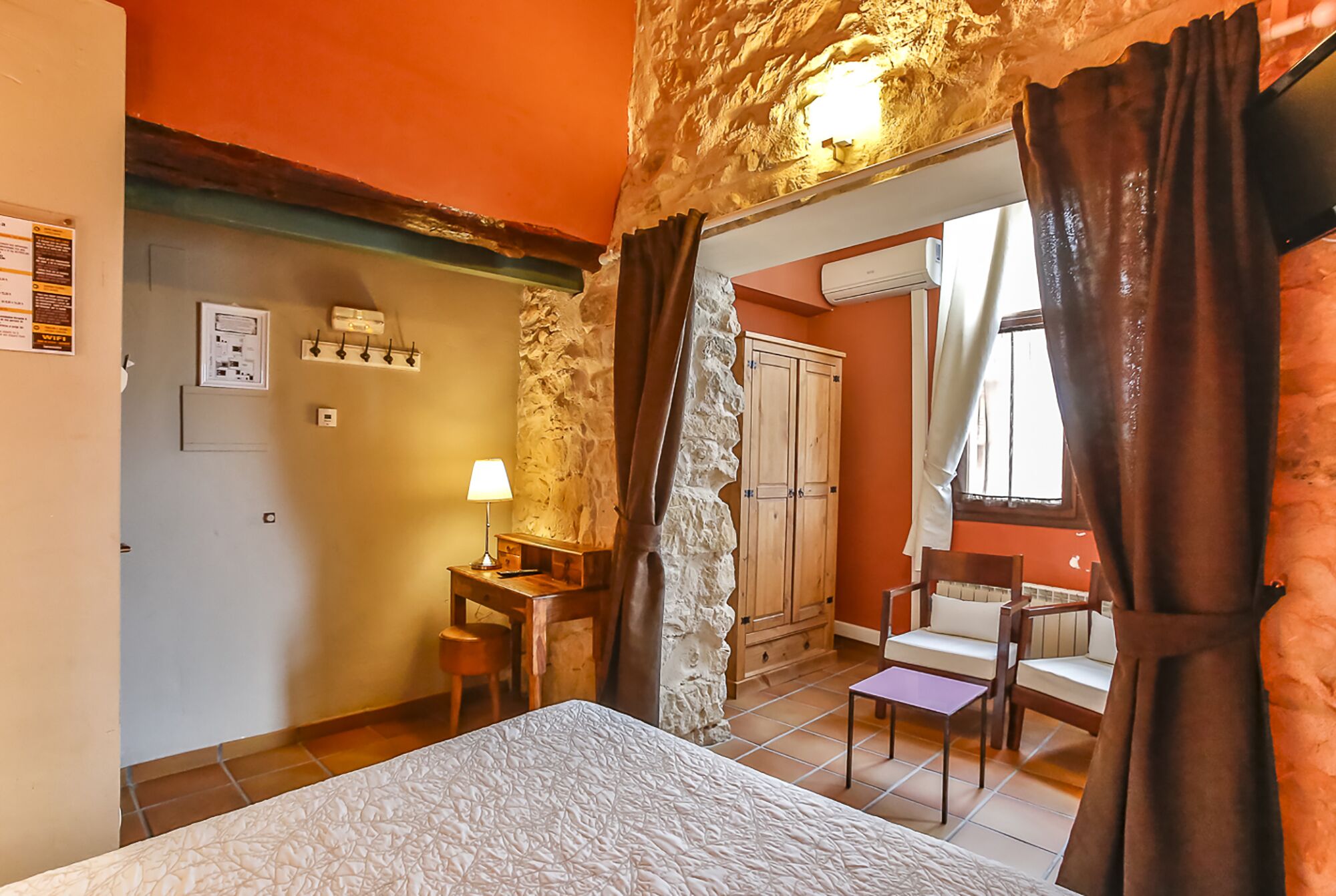 A bedroom with a bed, a desk with a lamp, a chair, a wardrobe, and a window with curtains.