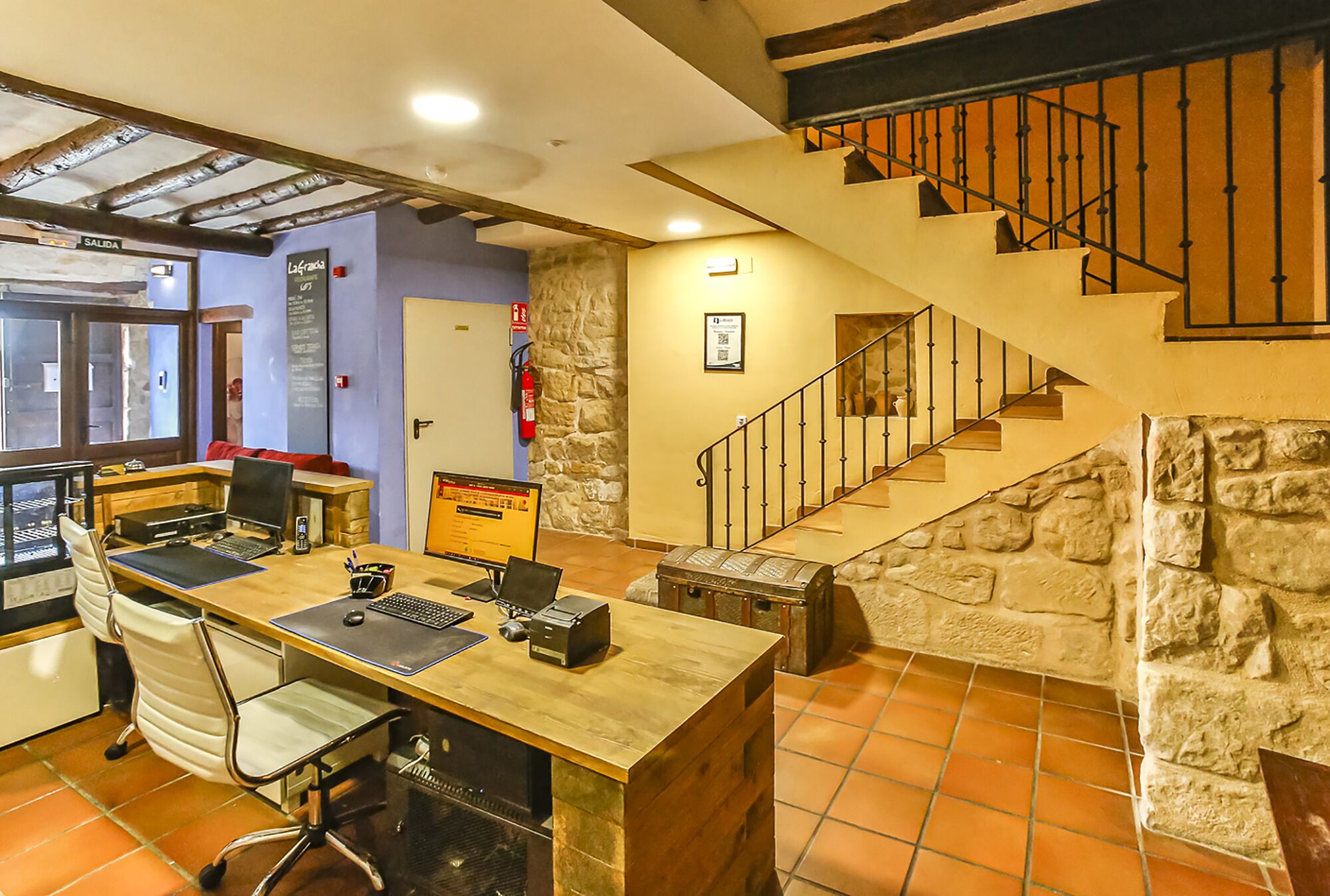 Un vestíbulo de hotel con un escritorio de madera, una computadora y una silla, una pared de piedra y una escalera.
